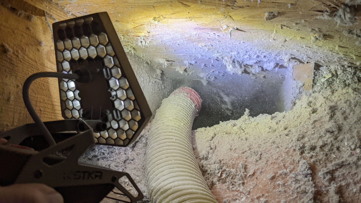 Large hose blowing attic insulation into a dark space lit up by a Mobile Task Light
